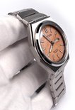 Zilverkleurig titanium horloge met een perzikkleurige wijzerplaat. De kast is vierkant met afgeronde hoeken.