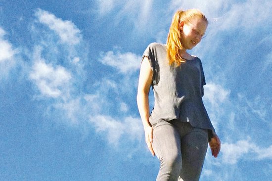 Jong meisje met blond haar in paardenstaart, lachend, in grijze kleding, tegen een blauwe lucht met wolken.