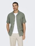 Model draagt groen, kortemouwige hemd met linnen textuur, wit T-shirt en beige broek.