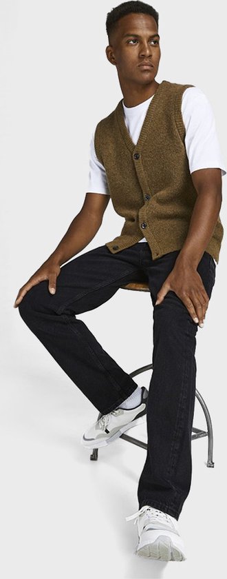 Man zittend op een krukje in een bruin gebreid vest, wit T-shirt en zwarte jeans.