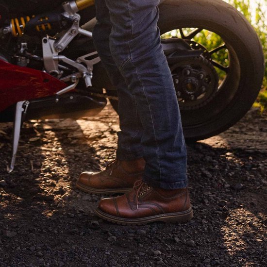 Mannen dragen bruine leren motorlaarzen met blauwe jeans naast een rode motor.