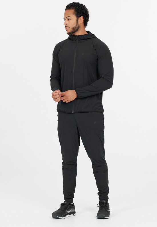 Man in een zwarte sportoutfit, bestaande uit een jas met capuchon en een trainingsbroek. Hij draagt sneakers.