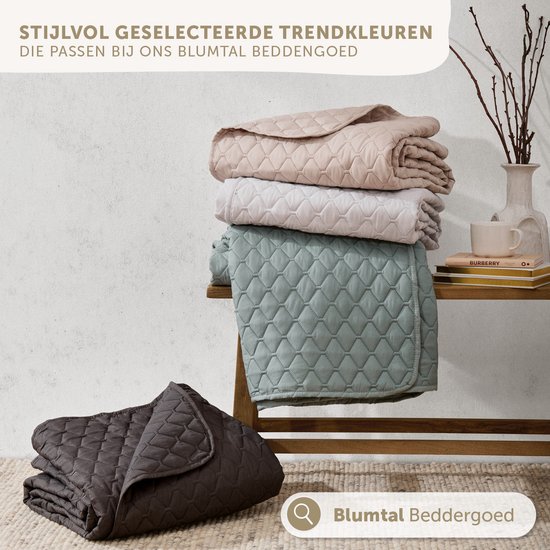 Gestapelde, gewatteerde spreien in beige, lichtgrijs en groen. Een donkergrijze sprei ligt vooraan.