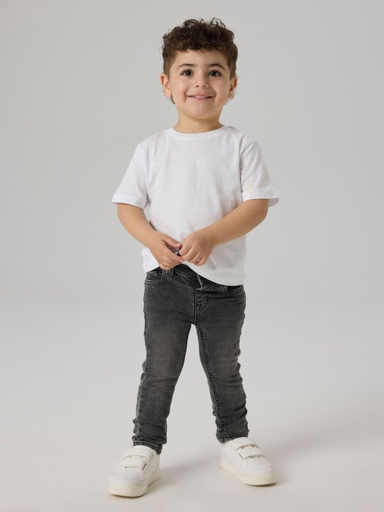 Jongen met krullend haar in een witte T-shirt en donkergrijze spijkerbroek.