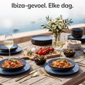 Een gedekte tafel met een Ibiza-gevoel, met blauwe borden, schalen met pasta en olijven, wijnglazen en bloemen.