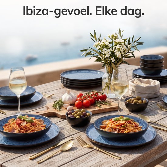 Een gedekte tafel met een Ibiza-gevoel, met blauwe borden, schalen met pasta en olijven, wijnglazen en bloemen.