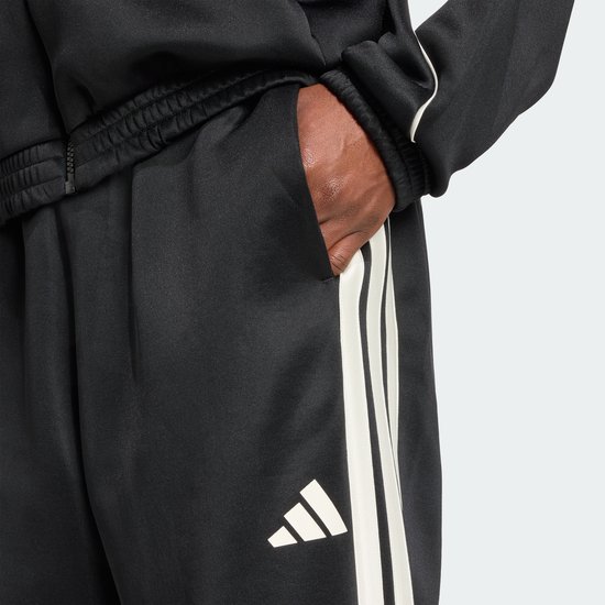 Detail van de zak van een zwarte trainingsbroek met witte strepen en Adidas logo.