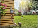 Gardena besproeiingscomputer, aangesloten op een kraan, met sproeiende sprinkler in de tuin.