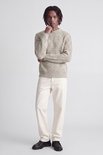 Model met dreadlocks draagt een beige trui met witte broek, hand bij de kin.