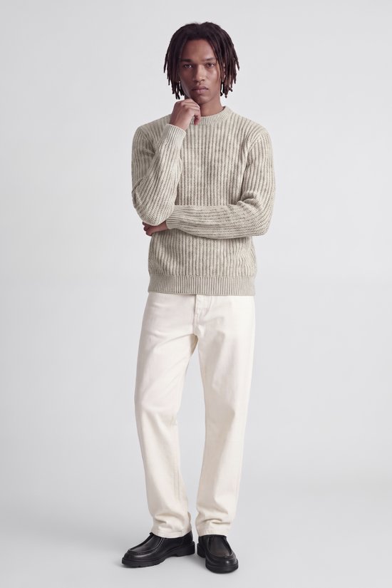 Model met dreadlocks draagt een beige trui met witte broek, hand bij de kin.
