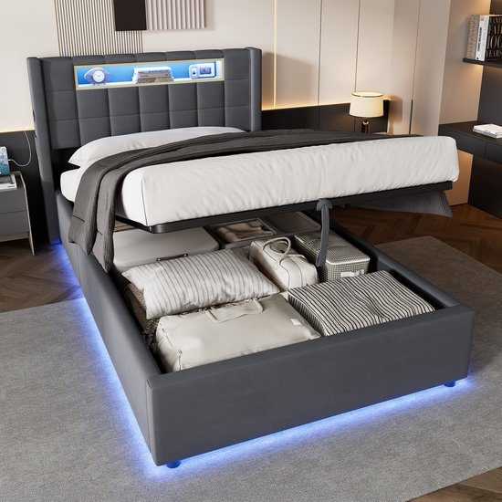 Donkergrijs bed met opklapbare opbergruimte onder de lattenbodem, gevuld met kussens en dekens.