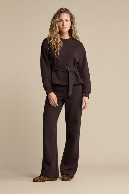 Model met blonde krullen in een bruine sweater en bijpassende broek, met een riem om de taille.