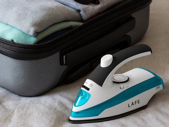 Wit en turquoise Lafe stoomstrijkijzer met zwarte handgreep, naast een koffer met kleding.