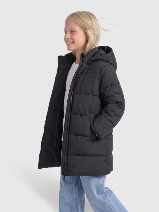 Blond meisje draagt een zwarte gewatteerde jas met capuchon over een wit T-shirt en lichtblauwe jeans.