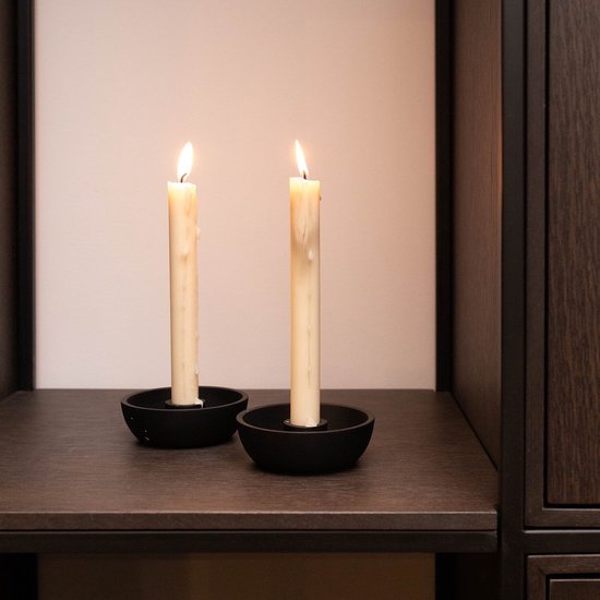 Twee brandende dinerkaarsen in zwarte metalen houders, geplaatst op een donkere plank in een interieur.