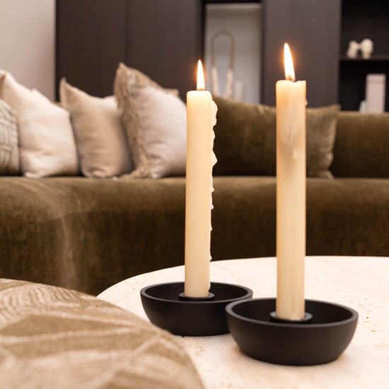 Twee brandende dinerkaarsen in zwarte houders op een lichte ronde tafel in een sfeervolle woonkamer.