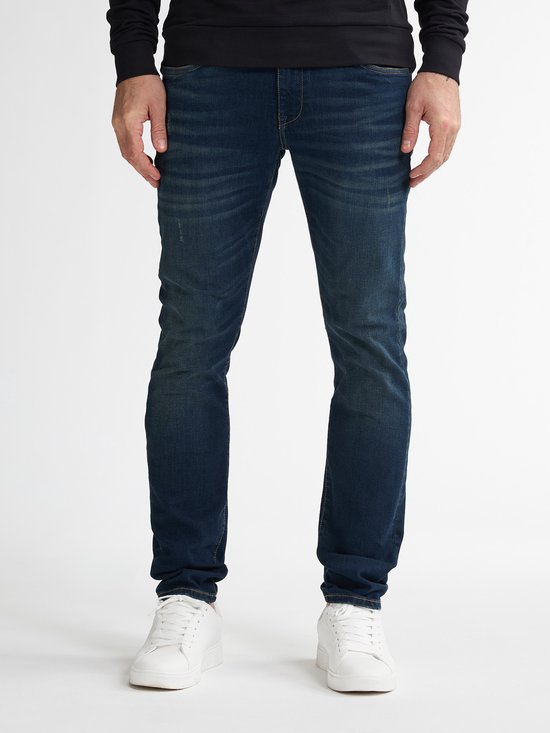 Man in donkerblauwe slim fit jeans met witte sneakers, voorkant benen.