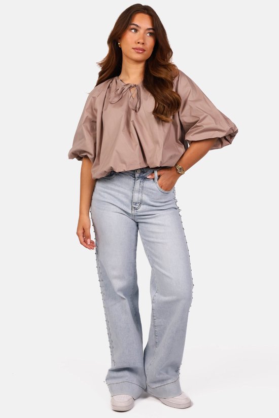 Model draagt een taupe ballon-top met pofmouwen en strikdetail, gecombineerd met lichtblauwe jeans met studs.