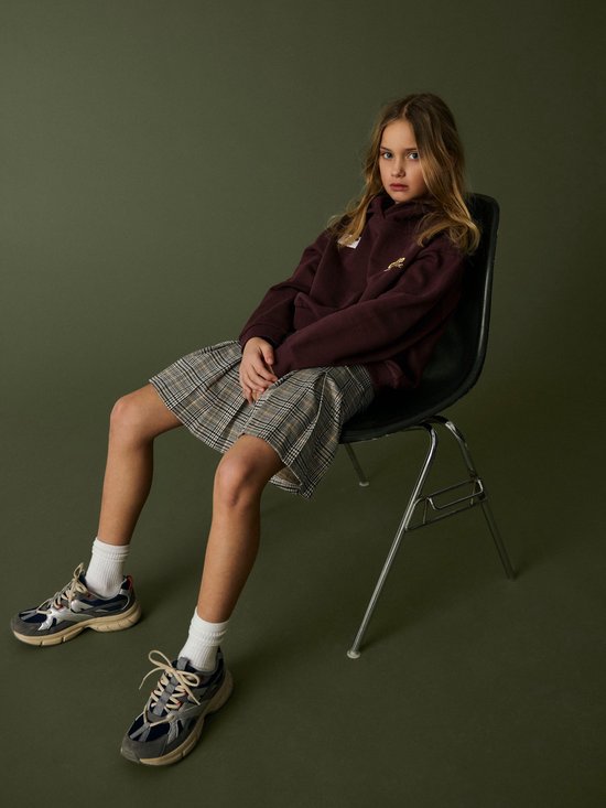 Model zittend in een bordeauxrode hoodie, geruite rok, witte sokken en sneakers tegen een groene achtergrond.