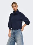 Model draagt een marineblauwe coltrui en een spijkerbroek met metalen studs.