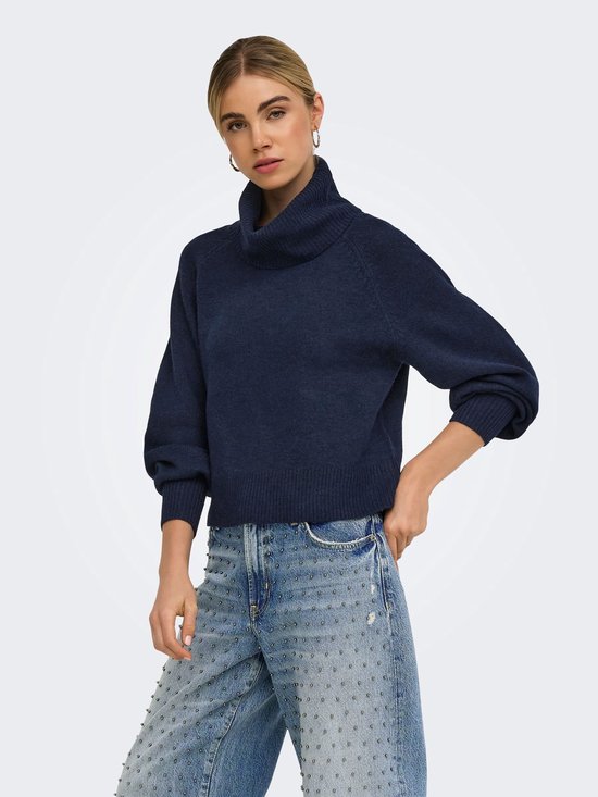 Model draagt een marineblauwe coltrui en een spijkerbroek met metalen studs.