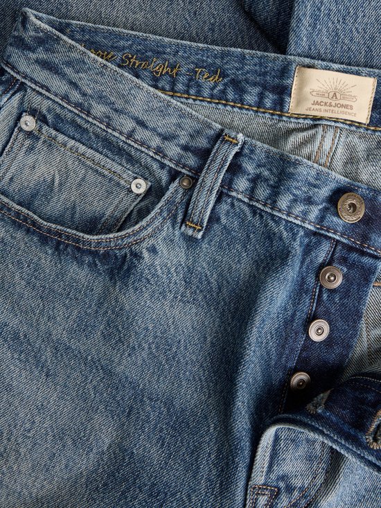 Detail van de voorkant van een spijkerbroek met knopen, zakken en een label met 'JACK & JONES JEANS INTELLIGENCE'.