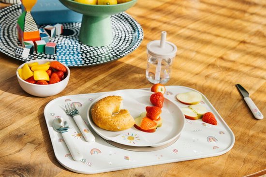 Service pour enfants avec imprimé soleil et arc-en-ciel sur plateau blanc, avec fruits, bagel et tasse.