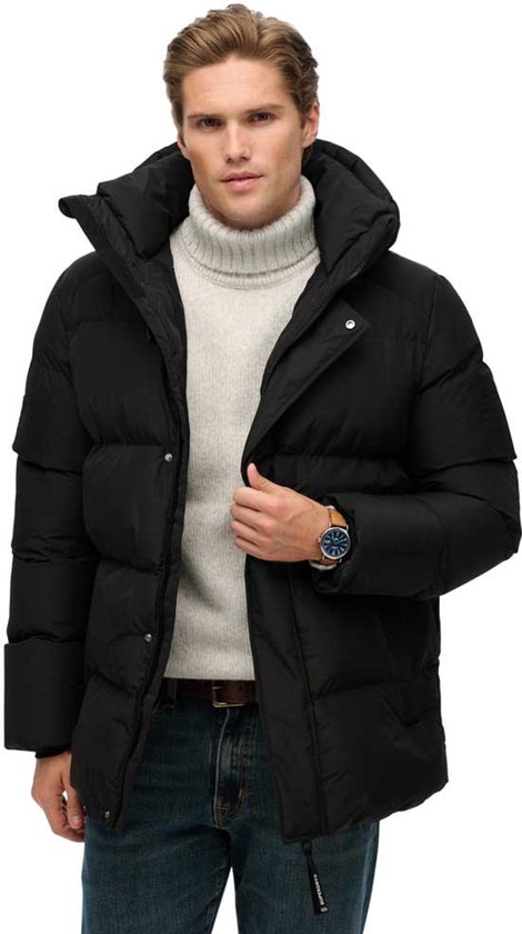 Zwarte Superdry pufferjas met capuchon, gedragen door een man met een grijze coltrui, spijkerbroek en horloge.