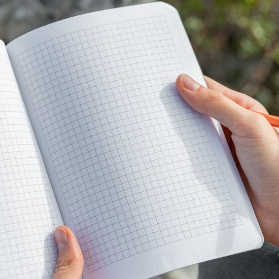 Carnet ouvert gris quadrillé, tenu par une personne avec un crayon orange.