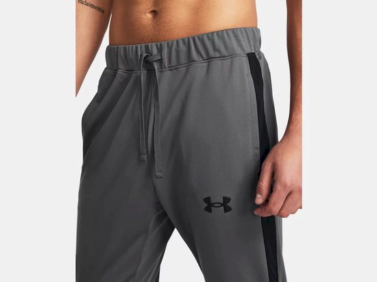 Detail van de broek van een grijs Under Armour trainingspak. Elastische tailleband met trekkoord, zwarte streep en logo.