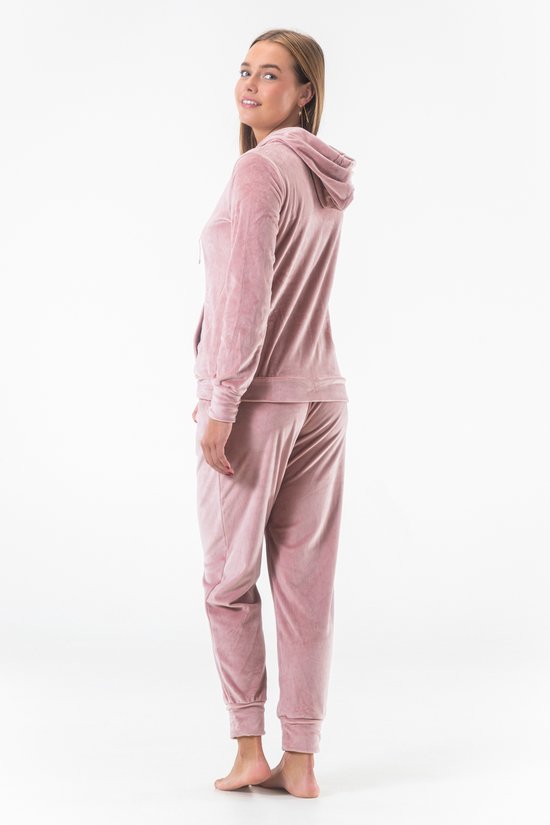Model in een lichtroze fluwelen joggingpak, bestaande uit een hoodie met rits en een joggingbroek. Ze draagt het pak van de zijkant, met haar rug naar de camera.