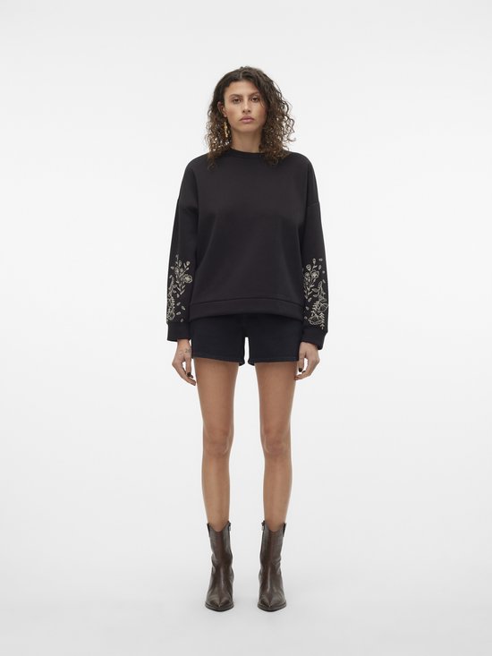 Model in zwarte denim shorts en een zwarte sweater met geborduurde bloemen op de mouwen.