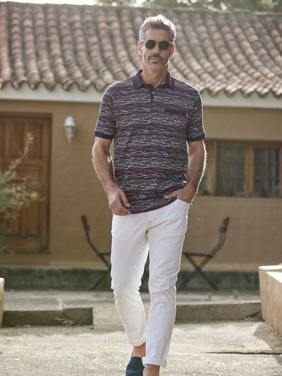 Man in een donkerblauwe polo met golvend streepjespatroon, beige broek en blauwe instappers, buiten.