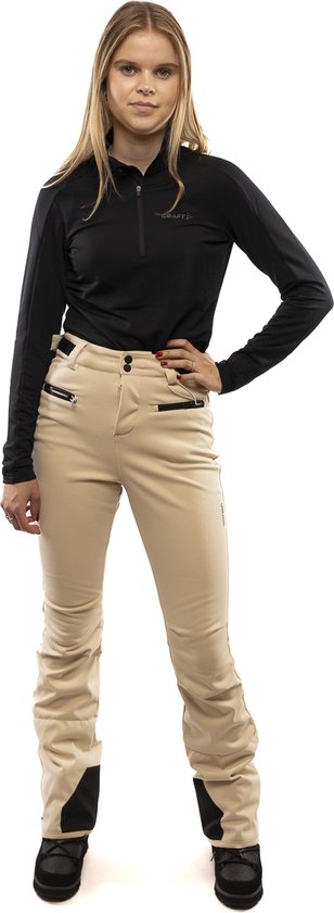 Vrouw die een beige skibroek met zwarte top en ski-uitrusting draagt.