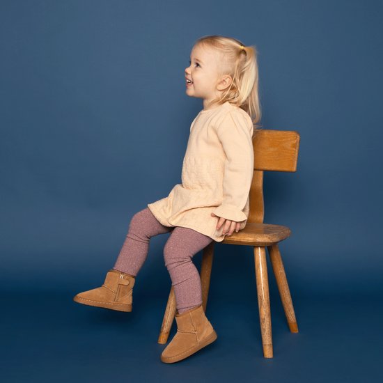 Een meisje met blond haar in een staartje zit op een houten stoel. Ze draagt een lichtroze jurk, glinsterende roze panty's en bruine laarsjes.