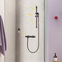 Zwarte thermostaatkraan in een douchecabine met iconen voor temperatuurbeveiliging en waterbesparing. Een paarse handdoek hangt links.