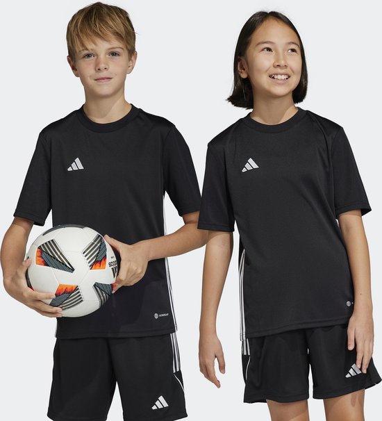 Kinderen in zwarte adidas Tabela 23 voetbalshirts en shorts, een jongen met voetbal, een meisje kijkt weg.