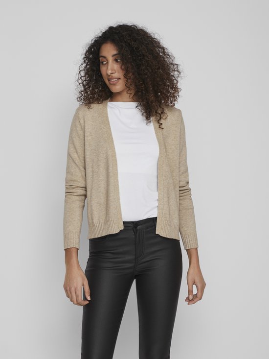 Lichtbeige gebreid vest met lange mouwen, gedragen over een wit shirt en zwarte leren broek.