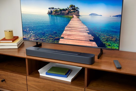 Barre de son JBL noire avec télécommande sur un meuble TV en bois, sous une grande télévision affichant un paysage.