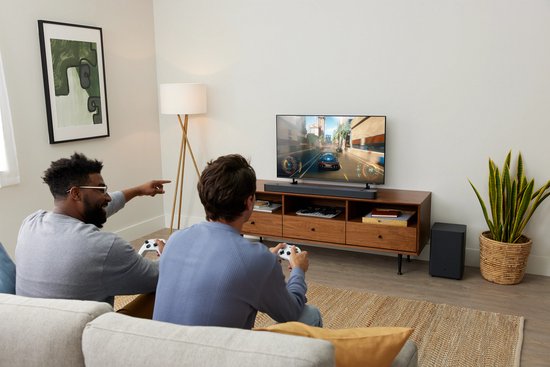 Twee mannen spelen een spel op tv, met de zwarte JBL soundbar en subwoofer in de woonkamer.