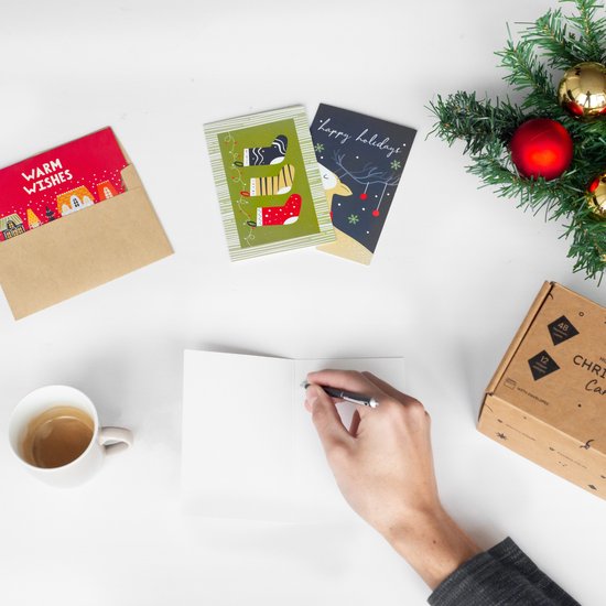 Bovenaanzicht van hand die kaart schrijft. Op tafel: kerstkaarten, envelop, koffiekopje, kerstdecoratie en opbergdoos.