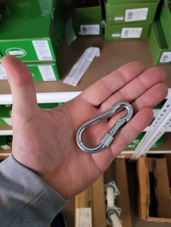 Een hand die een zilveren karabijnhaak met schroefsluiting vasthoudt. De achtergrond toont dozen en planken.