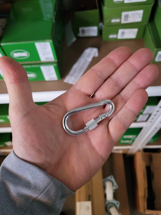 Zilverkleurige karabijnhaak met schroefsluiting in een handpalm, met dozen op de achtergrond.
