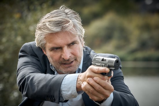 Man met grijs haar richt een pistool in de verte.
