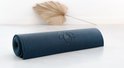 Opgerolde donkerblauwe yogamat met ribbelpatroon en subtiel lotuslogo, op een witte ondergrond met een gedroogde palmblad.