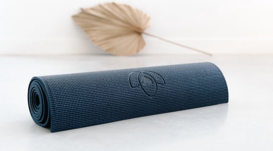Opgerolde donkerblauwe yogamat met ribbelpatroon en subtiel lotuslogo, op een witte ondergrond met een gedroogde palmblad.