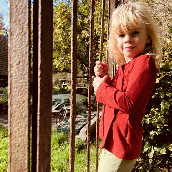 Meisje met blond haar in een rood wikkelvestje en lichtgroene broek, staand bij een roestig hek.
