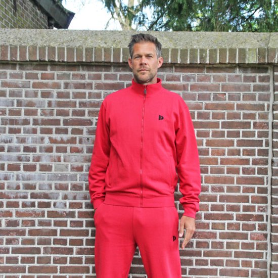 Man met donker haar draagt een rood trainingsvest en broek, staand voor een bakstenen muur.
