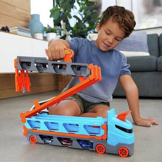 Jongen op de vloer die een blauwe Hot Wheels trailer met oranje oprijplaten en grijze opslagbaan speelt.