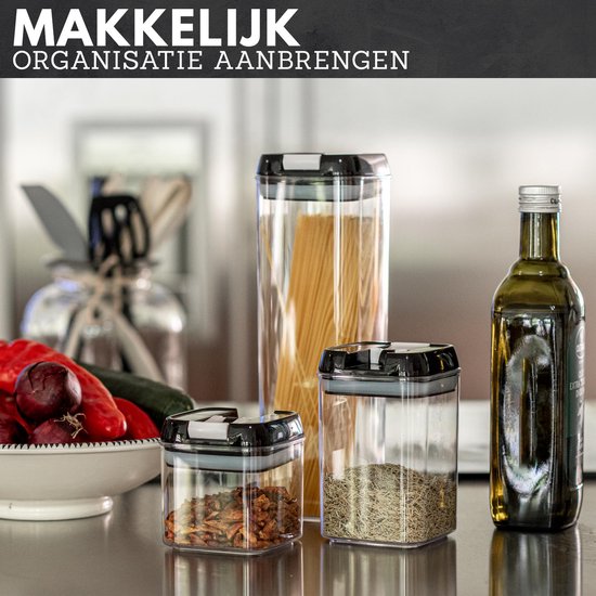 Set van doorzichtige voorraadpotten met zwarte deksels, gevuld met spaghetti, kruiden en gedroogde vruchten, naast verse groenten en olijfolie.